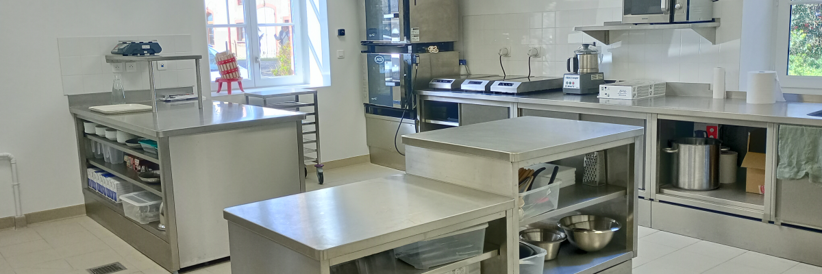 Cuisine professionnelle avec plans de travail, étagères, évier, réfrigérateur, micro-ondes, hotte et divers équipements.