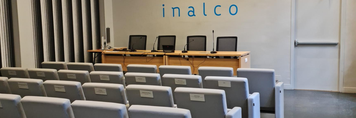 Salle de classe avec rangées de chaises, bureau, ordinateurs et tableau.