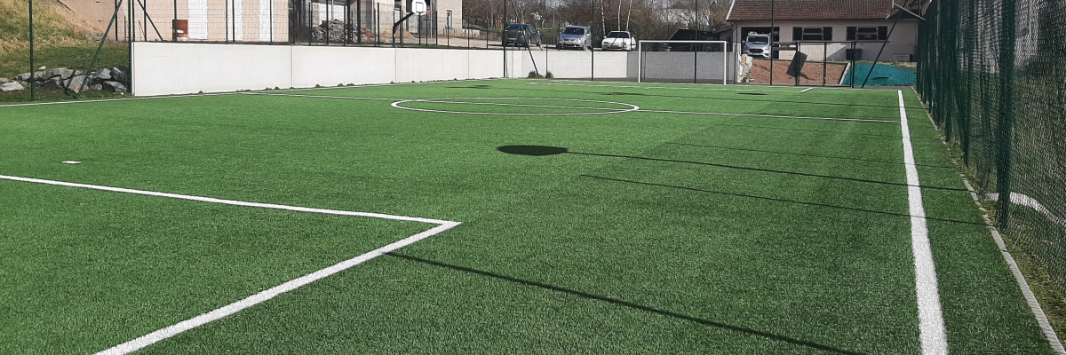 Terrain de sport avec gazon synthétique, panier de basket et clôture.