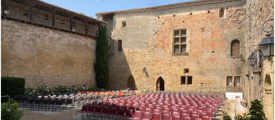 Cour de château avec mur en pierre, fenêtres, porte, chaises rouges, lampadaire et végétation.