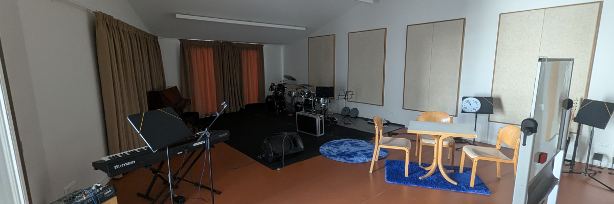Studio avec clavier, batterie, micros, table, chaises, ampli et matériel audio.