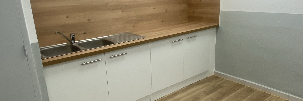 Cuisine avec plan de travail, évier, meubles blancs, sol en bois, climatiseur et fenêtre.