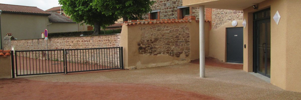 Cour avec sol en terre battue, bâtiment en pierre et briques, portail métallique, arbre.