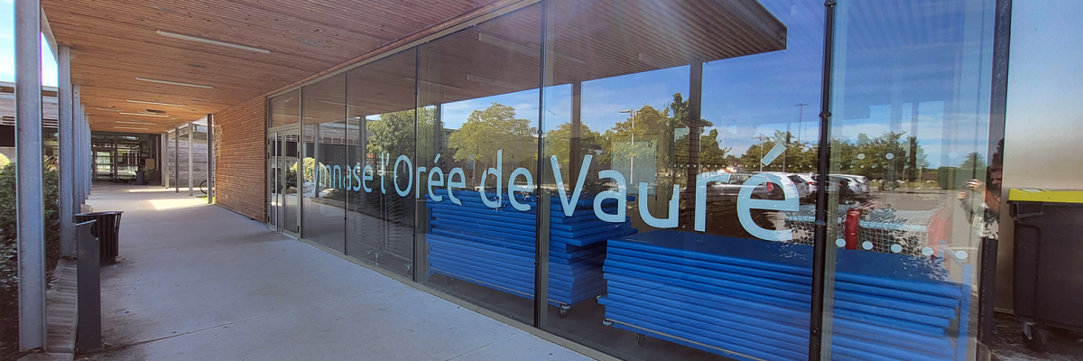 Entrée avec toit, murs vitrés, bancs à l'intérieur, arbres et ciel visibles à travers les vitres.