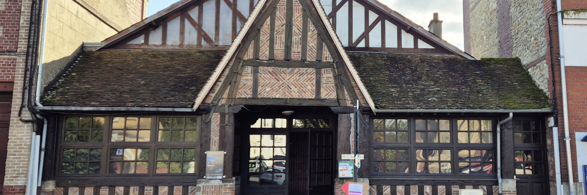 Bâtiment à structure en bois avec toit en tuiles, grandes fenêtres et porte centrale. Deux bancs devant, plantes et panneaux.
