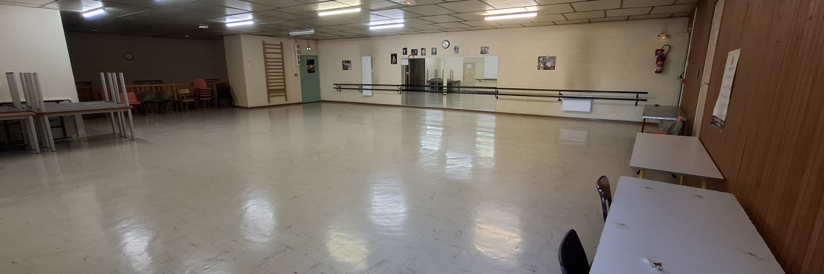Salle spacieuse avec tables, chaises, barres de danse, horloge, tableaux et éclairage au plafond.