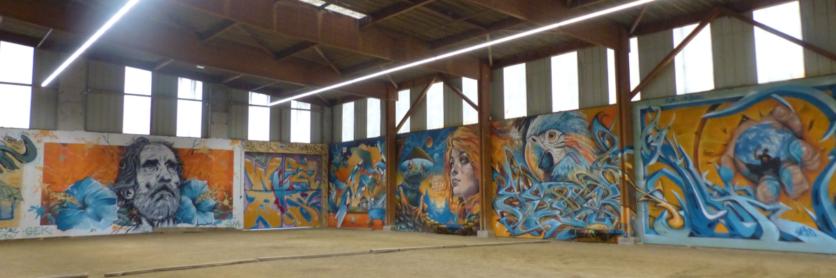 Atelier avec graffitis muraux, sol en béton, toit métallique, fenêtres.