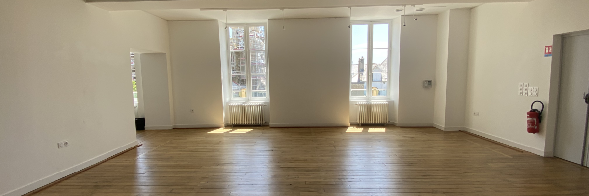 Salle vide avec plancher en bois, grandes fenêtres et porte rouge.