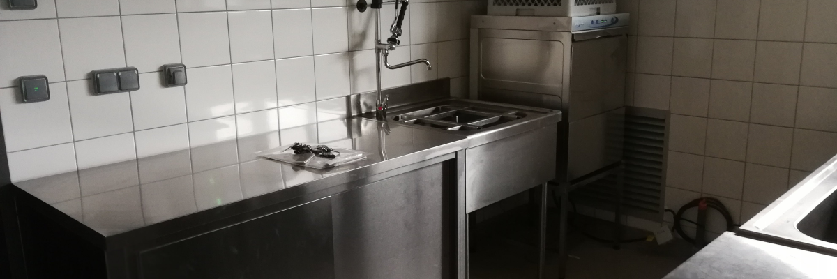 Cuisine avec évier en acier inoxydable, robinet à bras flexible, plaques de cuisson, friteuse et murs carrelés.