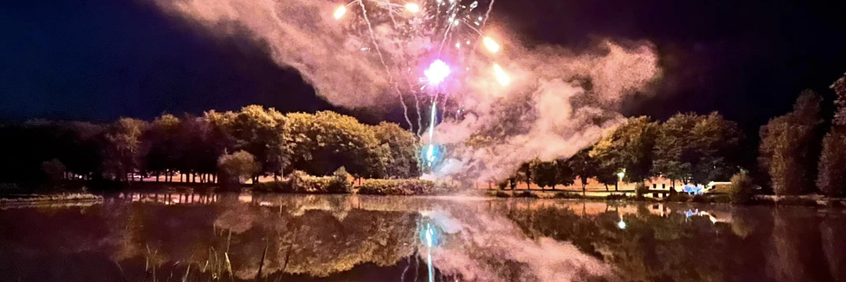 Feux d'artifice éclairant la nuit au-dessus d'un plan d'eau avec des arbres en arrière-plan.
