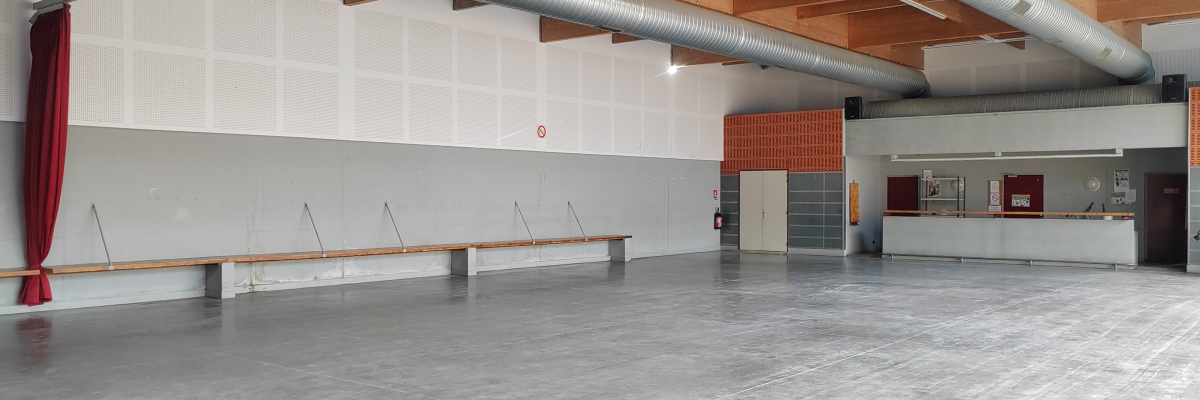 Salle vide avec bancs, tuyaux suspendus et mur partagé avec un gymnase.