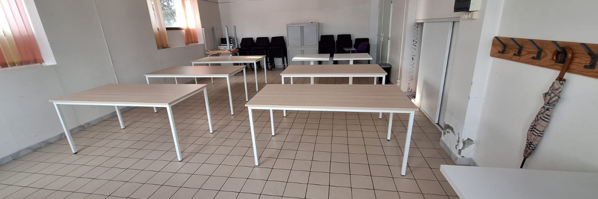 Salle avec tables, chaises, fenêtres, radiateur, et tableau d'affichage.