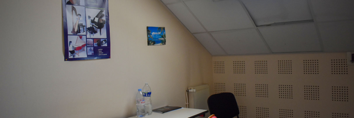 Petite pièce avec table, chaises, radiateur, bouteilles d'eau, et deux affiches sur le mur. Puits de lumière au plafond.