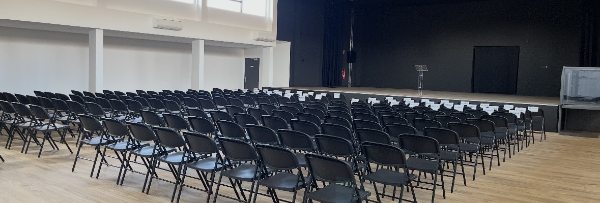Salle avec rangées de chaises, scène, écran et fenêtres.