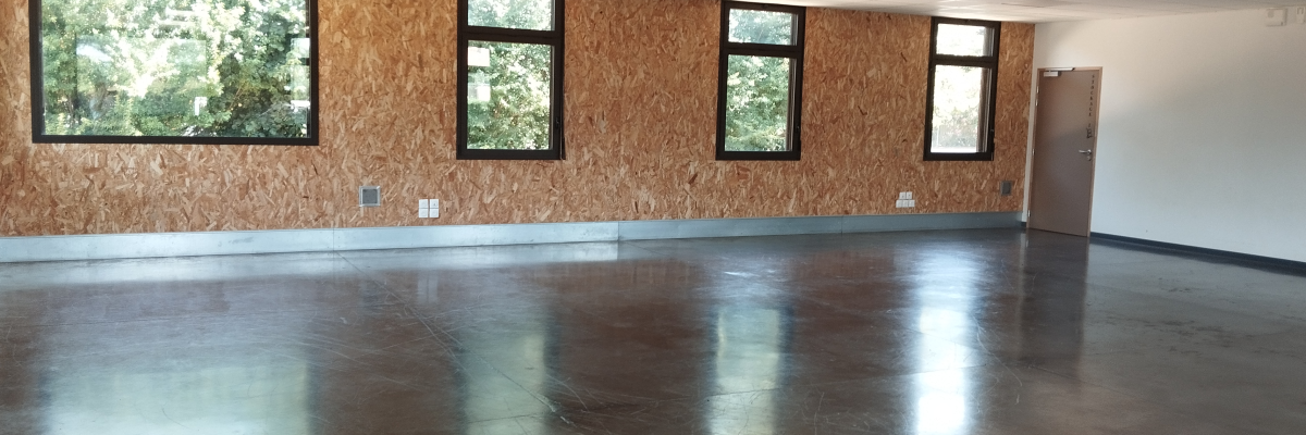 Salle vide avec sol poli, murs en OSB, fenêtres et porte.