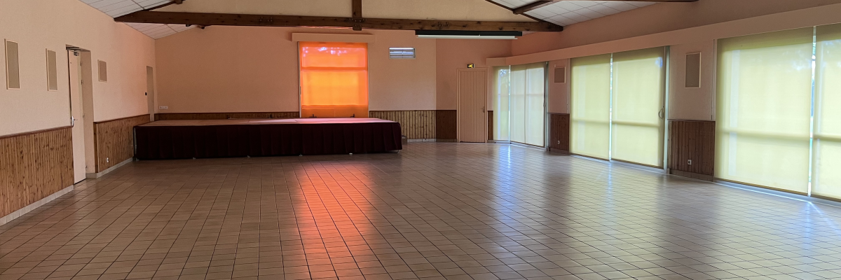 Salle spacieuse avec plancher en carreaux, scène, grandes fenêtres, portes et poutres apparentes.