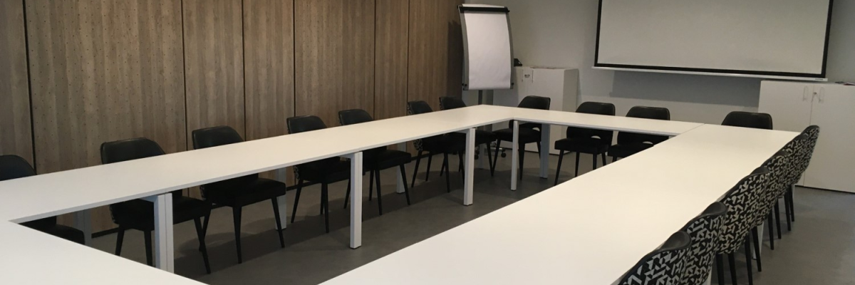 Salle de réunion avec table en U, chaises, écran blanc, et projecteur suspendu.