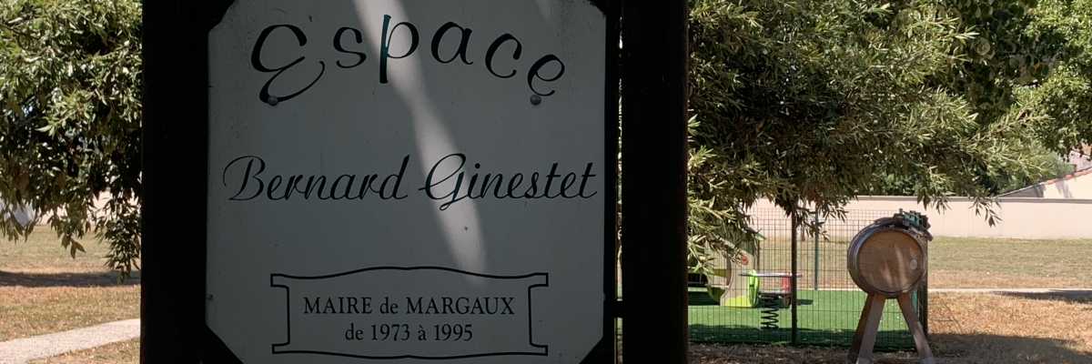 Cette image montre un panneau avec l'inscription "Espace Bernard Ginestet" et "Maire de Margaux de 1973 à 1995". À l'arrière-plan, il y a un cheval en statue, un arbre et un terrain herbeux.