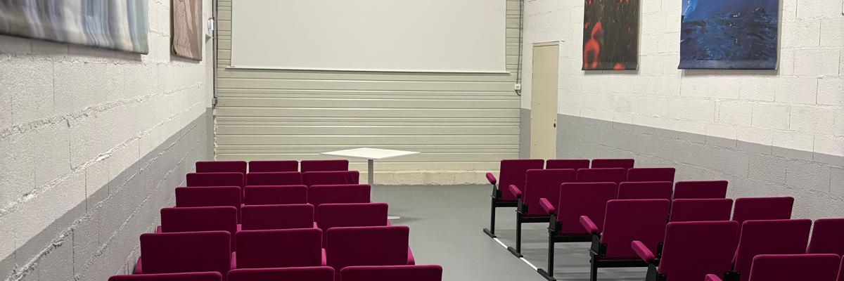 Salle avec rangées de chaises, tableaux aux murs, porte au fond.