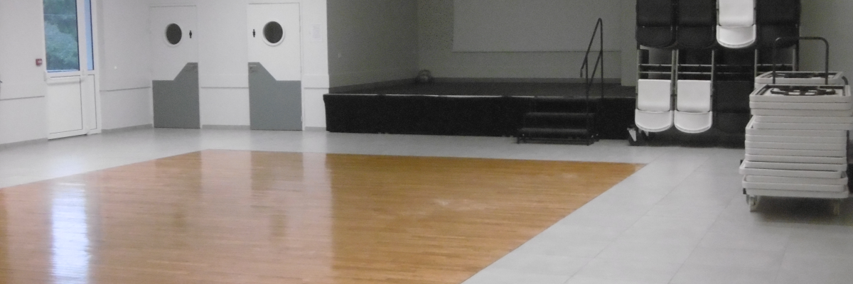 Salle avec parquet, portes, écran, chaises empilées et porte-manteaux.