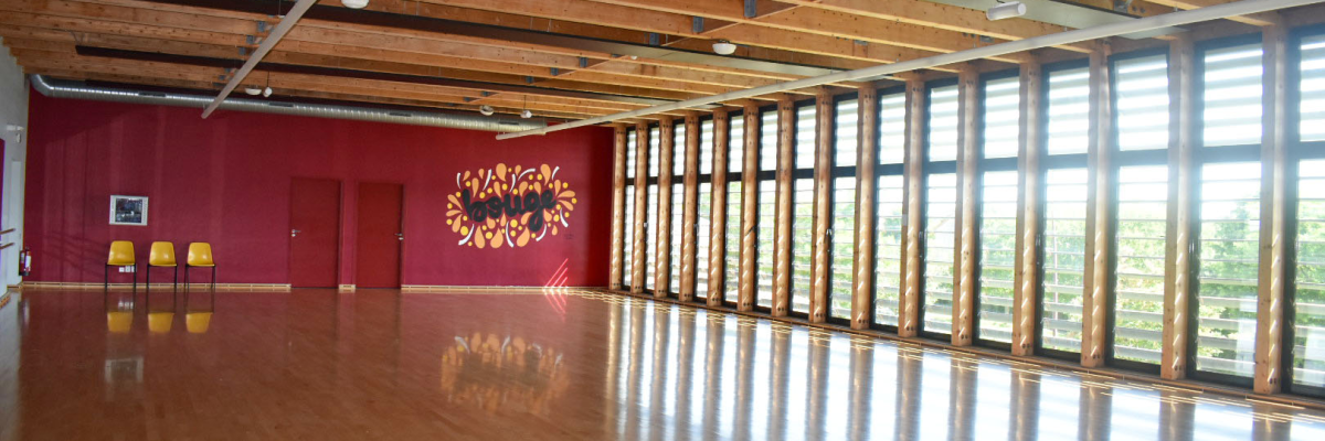 Salle spacieuse avec parquet, murs rouges, grandes fenêtres, et chaises proches du mur.
