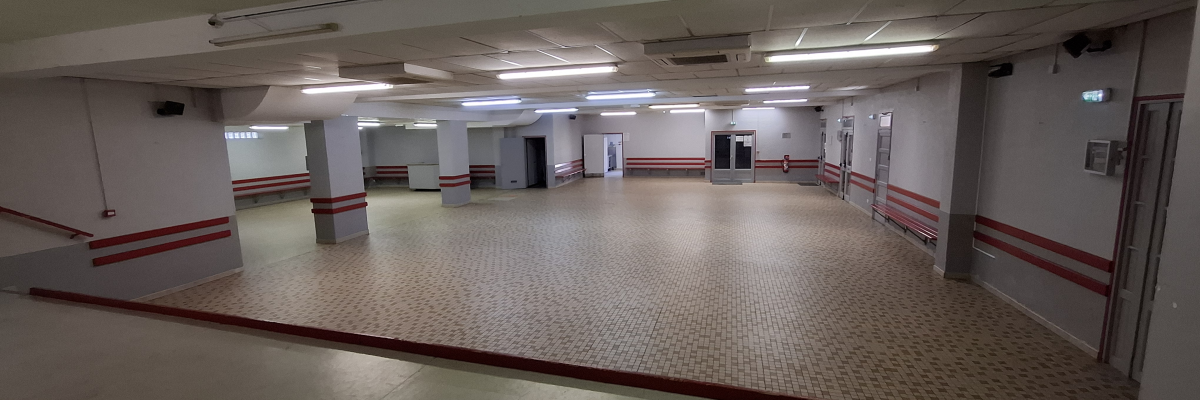 Grande salle vide avec éclairage au plafond, murs blancs avec bande rouge, sol carrelé.