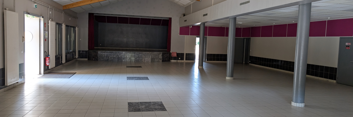 Salle vide avec toit en bois, sol carrelé, murs blancs et colonnes métalliques.