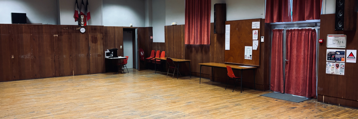 Salle avec parquet, rideaux rouges, tables, chaises, haut-parleurs, tableau d'affichage et porte.