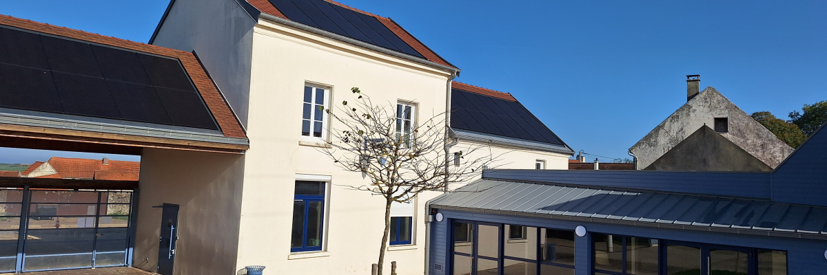 Bâtiment avec toits en pente, panneaux solaires, fenêtres, porte, arbre, poubelle, et extension moderne avec grandes vitres.