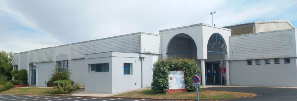 Bâtiment blanc avec entrées, fenêtres, buissons, parking et panneaux de signalisation.