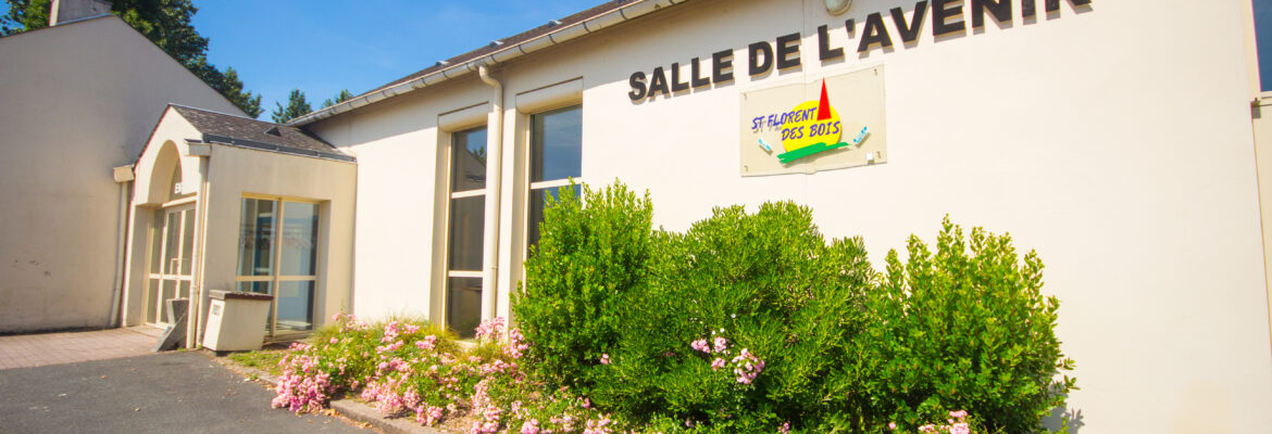 Bâtiment blanc avec enseigne "Salle de l'Avenir". Entrée avec porte vitrée. Buissons et fleurs.