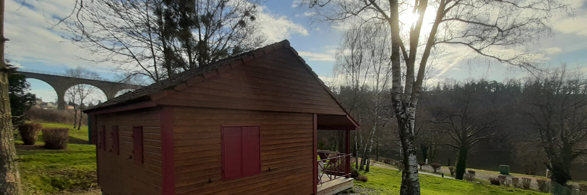 Cabane en bois avec volets rouges, terrasses, située dans un champ avec arbres et pont au loin.