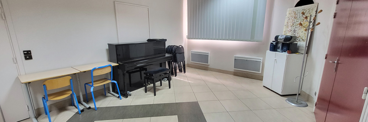 Salle avec piano, chaises, tables, tableau blanc, radiateurs, machine à café, porte-manteau.