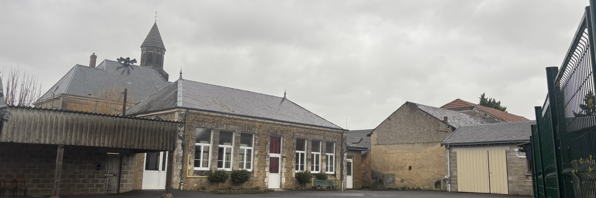 Bâtiment en pierre avec toit incliné, cour asphaltée, clôture métallique, abri de voiture.