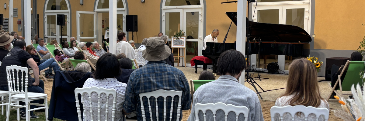 Concert en plein air avec piano, chaises, public et structure vitrée.