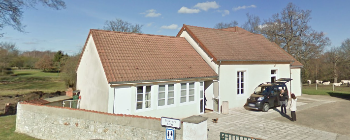 Maison blanche avec toit en tuiles rouges, voiture et personnes près d'une porte, mur de pierre et panneaux de signalisation.