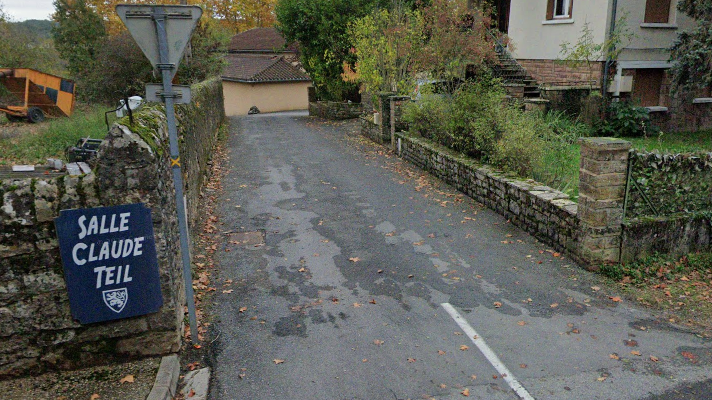 Rue étroite avec maison, panneaux de signalisation, réverbère et végétation.