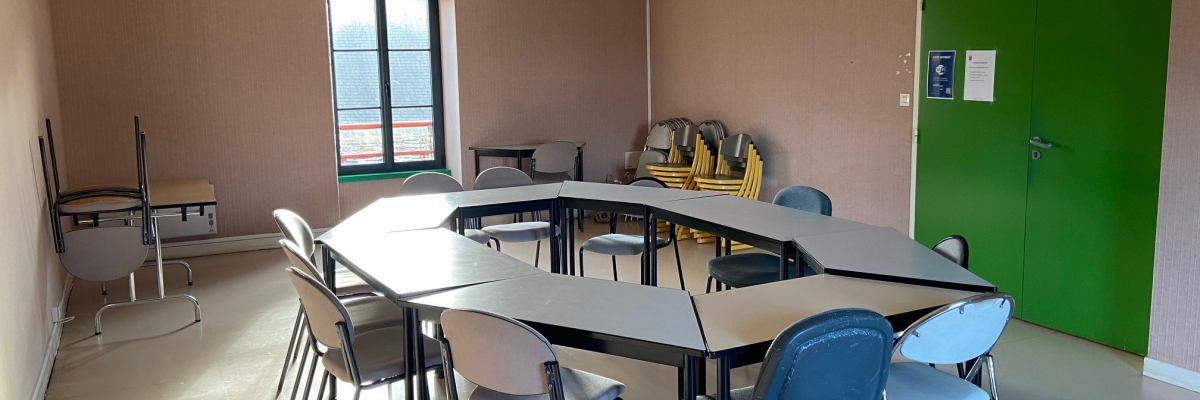 Salle avec table ovale, chaises, planches à dessin, porte verte et fenêtres.