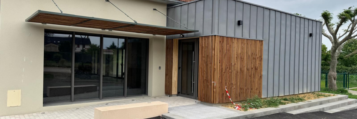 Bâtiment avec entrée vitrée, auvent en bois, porte en bois, banc et allée pavée.
