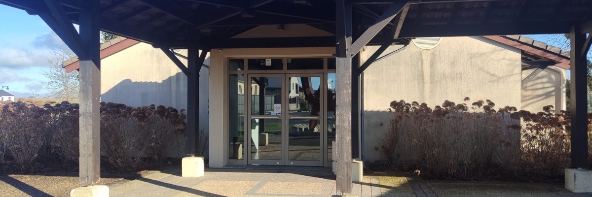 Entrée de bâtiment avec auvent, porte vitrée, allée et végétation.