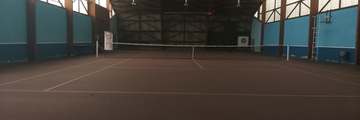 Terrain de tennis intérieur avec filet, murs bleus, toit métallique, échelle et panneau.