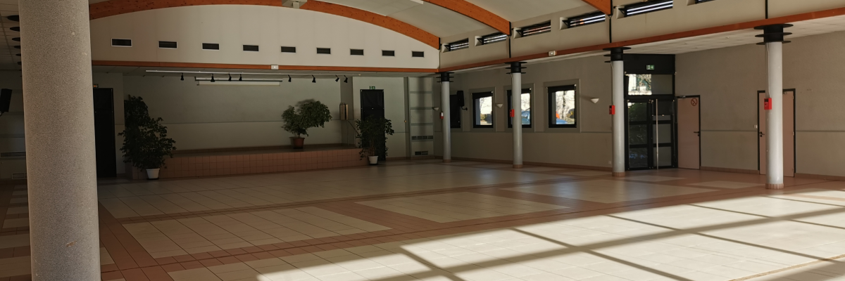 Grand espace intérieur avec colonnes, arches en bois, portes en verre, plantes et carrelage au sol.