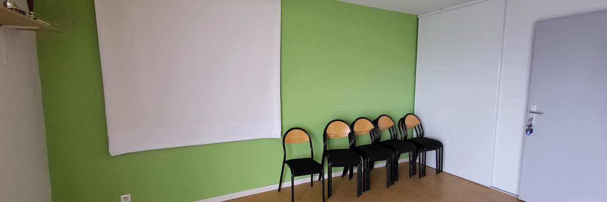 Salle avec mur vert, tablette, écran blanc, cinq chaises alignées, porte et sol bois.
