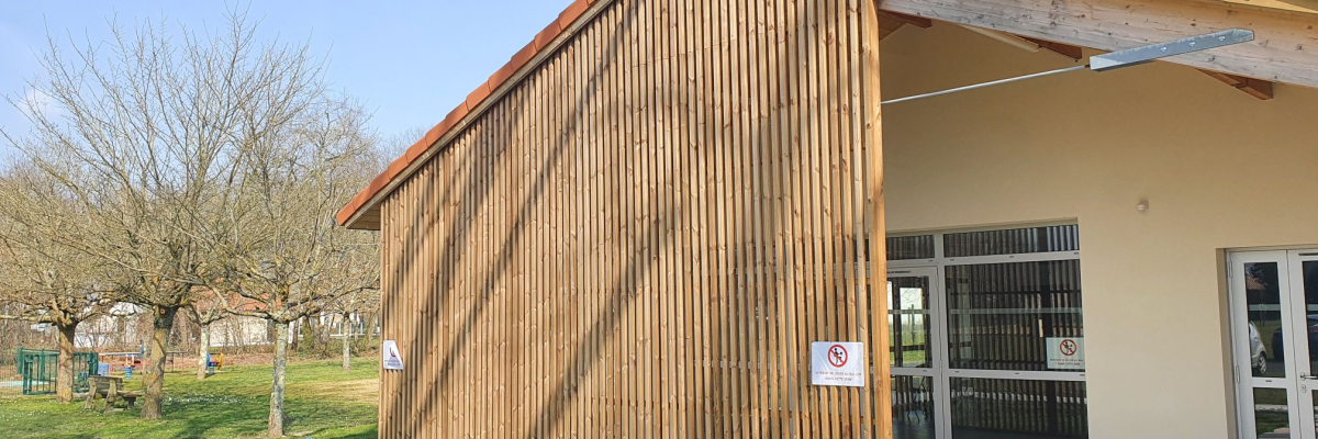 Bâtiment avec murs en bois et porte en verre, pelouse et arbres en arrière-plan.