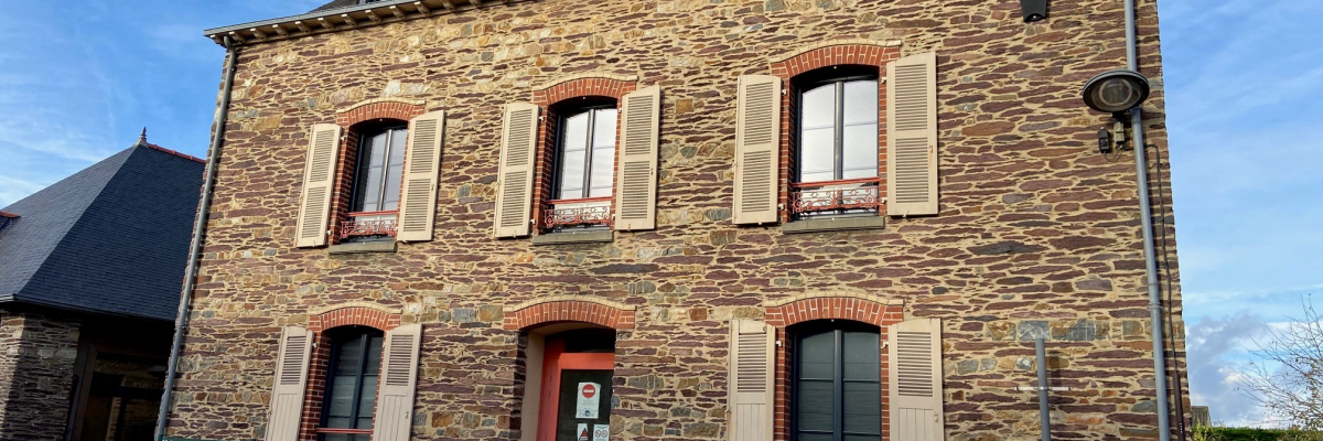 "Maison en pierre avec fenêtres à volets blancs, porte rouge, et toits mansardés."