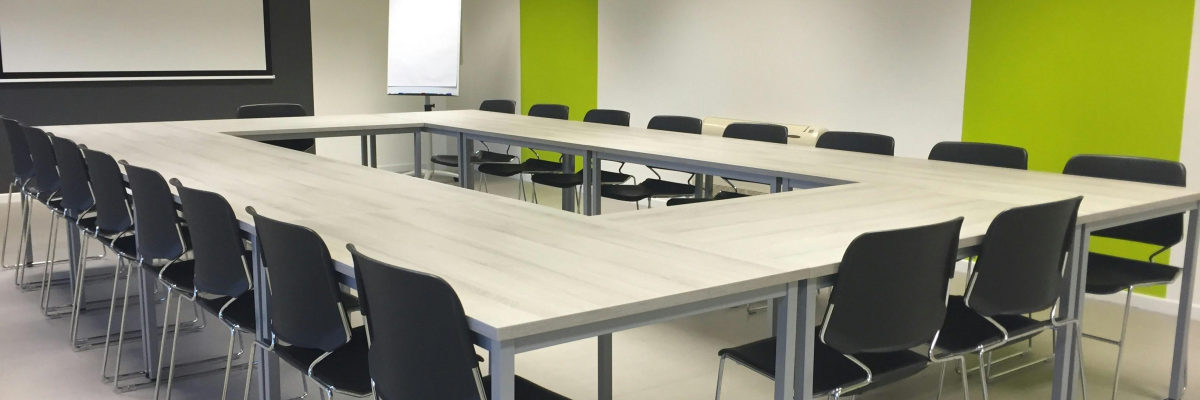 Salle avec table rectangulaire, chaises, écran, vidéo projecteur, luminasires et tableau blanc.