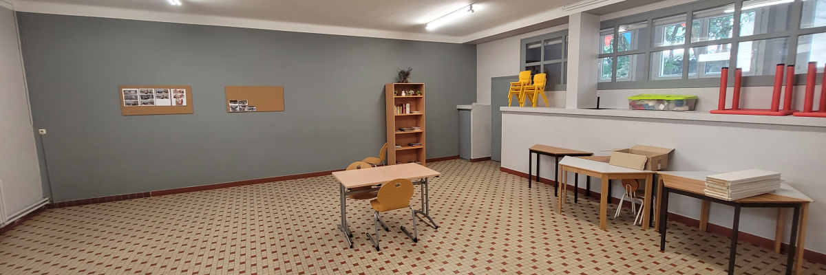 Salle de classe avec étagères, tables, chaises, bureau, plante et tableaux.