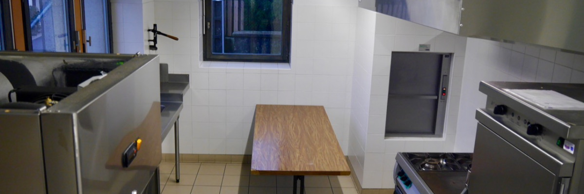 Cuisine avec table en bois, plan de travail, plaques de cuisson, hotte, évier et réfrigérateur.