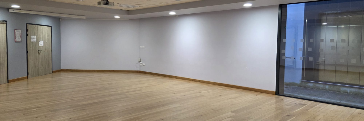 Grande salle vide avec parquet, plafonniers, projecteur, portes et fenêtres.