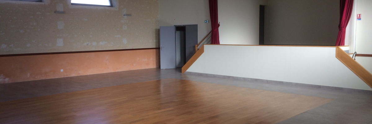 Salle vide avec parquet, murs beiges, plafond blanc, deux fenêtres rondes, escalier, rideau rouge.